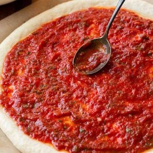 Um molho de tomate caseiro para pizza sendo colocado sobre uma massa de pizza com uma colher.